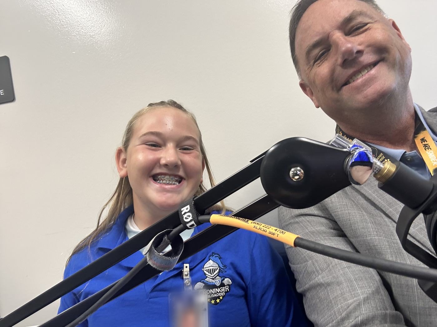 Noah Smith and a Croninger Elementary student in a Crusaders polo, both grinning behind two studio microphones on boom arms.