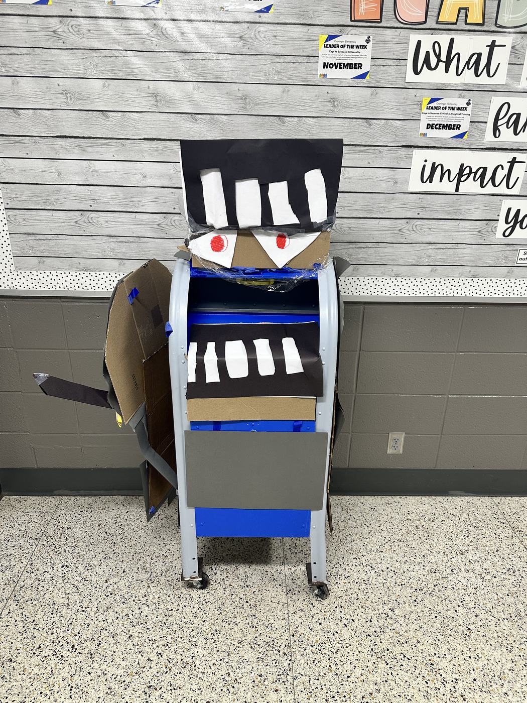 A cart decorated with cardboard and paper to look like a monster with large teeth and red eyes, parked against a classroom wall.