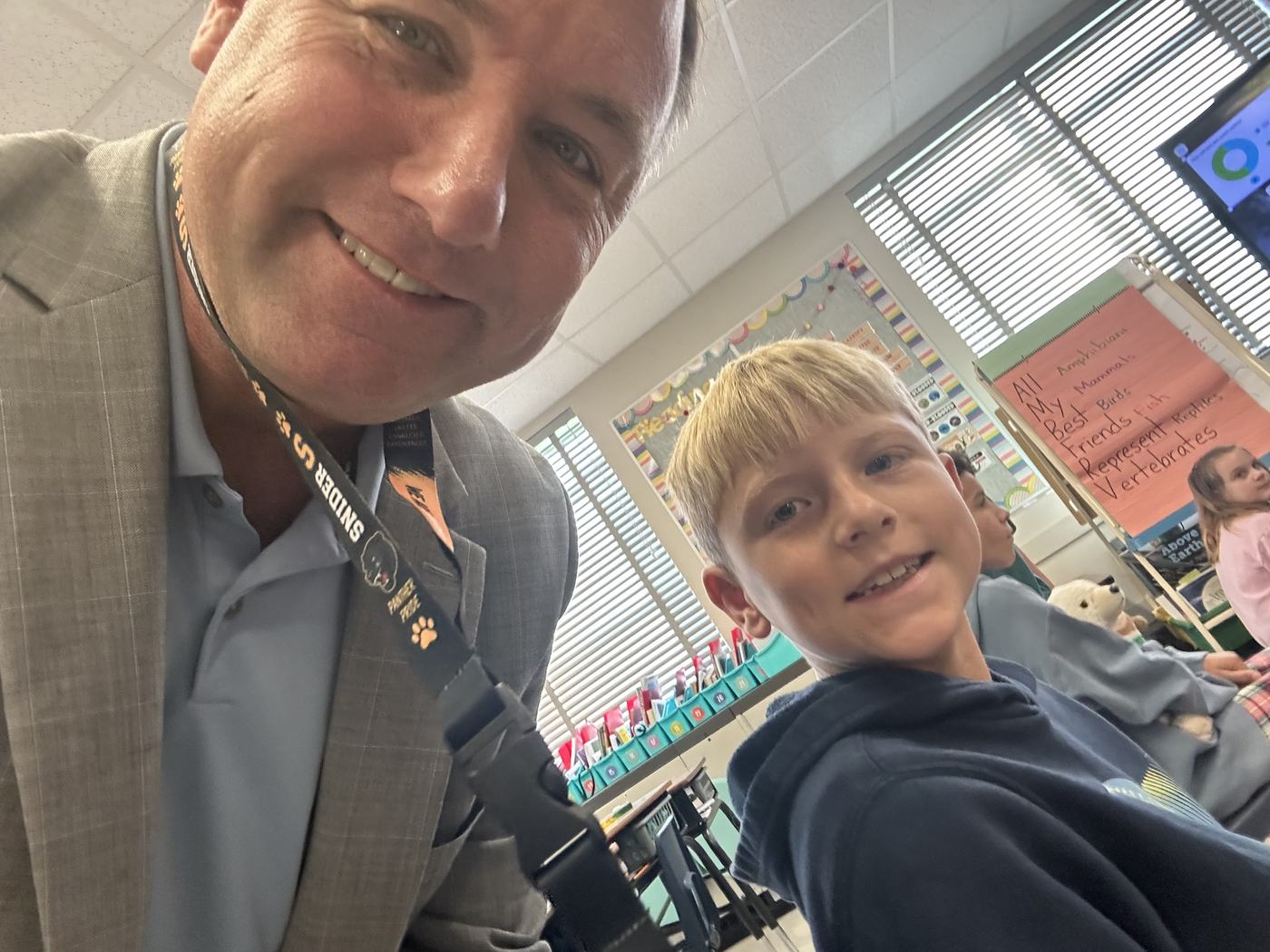 Noah Smith and a Croninger Elementary student in a classroom, a hand-lettered poster reading “All My Best Friends Represent Vertebrates” on the wall behind them.