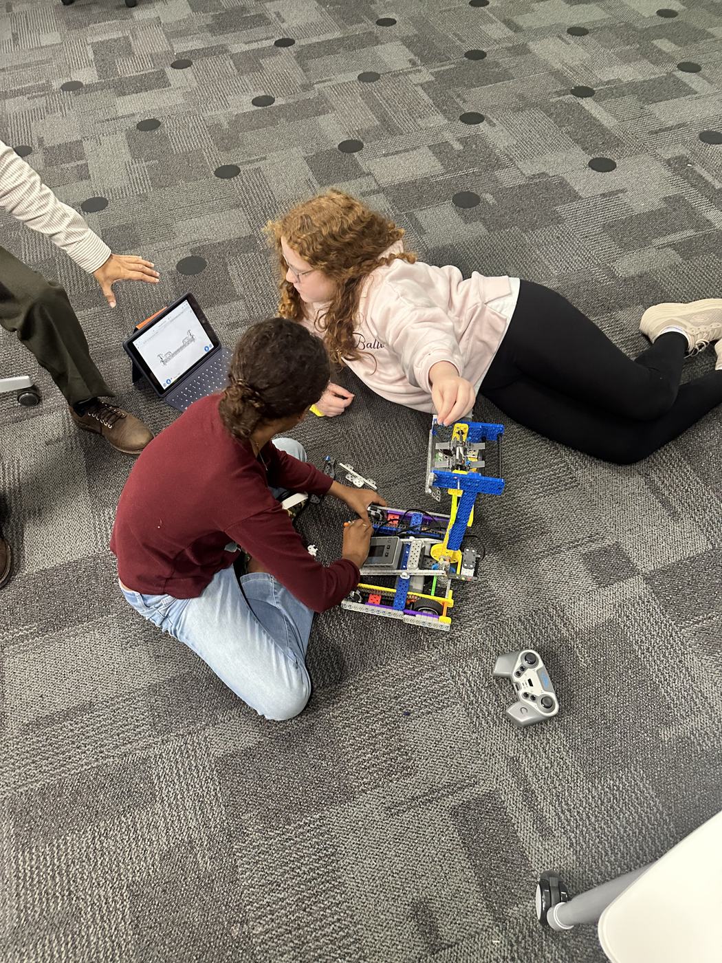Two students assembling a blue and yellow motorised robot on the floor, with a tablet open beside them showing their program.
