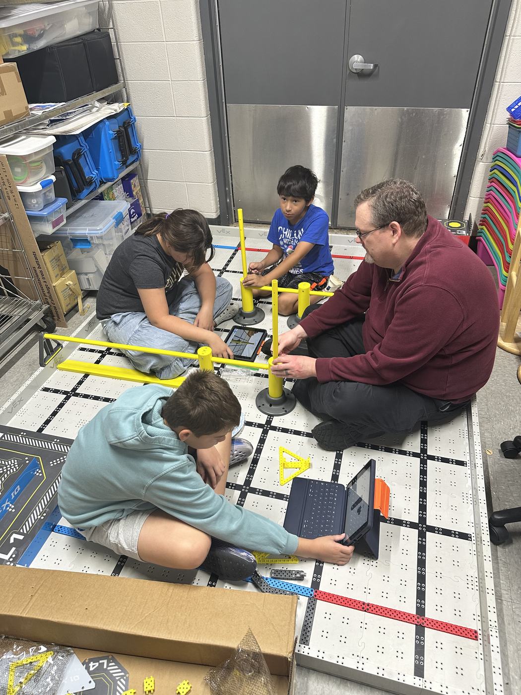 Three Croninger Elementary students and a coach sitting around a robotics practice mat, working on tablets and laptops.